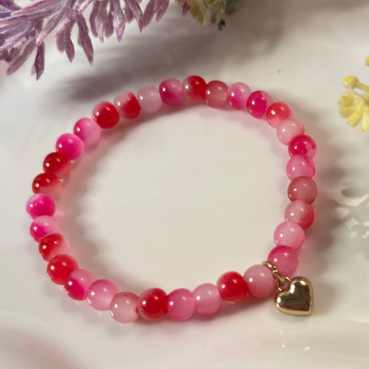 Pink beaded bracelet with a gold heart charm on a white surface with flowers.
