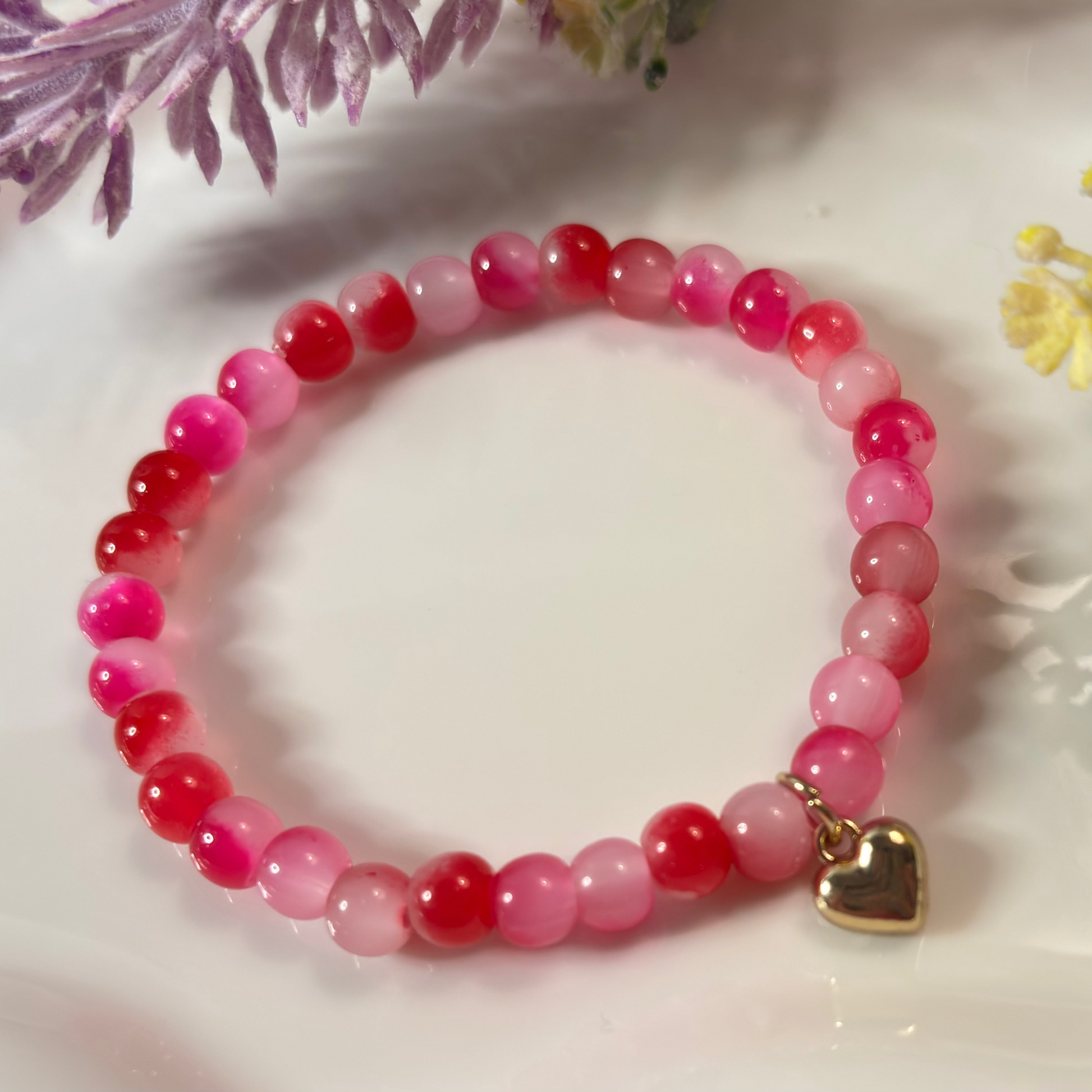 Pink beaded bracelet with a gold heart charm on a white surface with flowers.